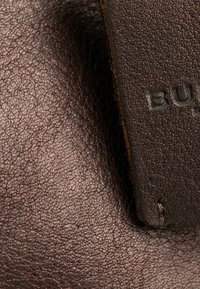 Surface en cuir brun avec une texture granuleuse, présentant une étiquette brune embossée avec "BURBERRY." Coutures visibles sur le bord.