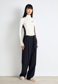 Maglietta sportiva a maniche lunghe bianca e nera con zip e logo "BOSS", abbinata a pantaloni larghi neri e sneakers con dettagli beige.