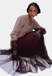 Kobieta w beżowym swetrze z biżuteryjnym dekoltem, ciemnej prześwitującej spódnicy, długich brązowych skórzanych rękawiczkach i wiszących kolczykach, siedząca na podłodze.