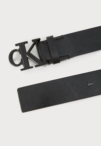 Ceinture en cuir noir avec une grande boucle en métal noir ornée des lettres "cK". La ceinture présente une texture lisse et des détails de couture minimaux.