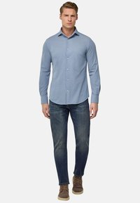 Camicia azzurra a maniche lunghe con colletto classico, abbinata a jeans scuri e scarpe in suede marroni, in piedi contro uno sfondo bianco.