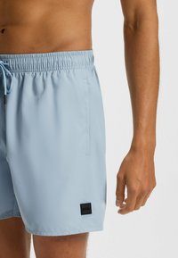 Pantaloni da bagno azzurri in tessuto leggero, con vita elasticizzata e cordoncino, tasche laterali e una piccola etichetta nera logo.