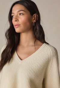 Krémový rebrovaný sveter s výstrihom do V, voľnými rukávmi a jemnou textúrou, doplnený neutrálnym pozadím.
