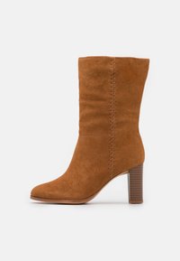 Bota de gamuza color tostado, hasta la pantorrilla, con tacón apilado y costura vertical decorativa a lo largo del costado, sobre un fondo blanco.