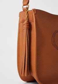 Borsa a mano in pelle marrone con finitura testurizzata, dotata di un accento con nappina, hardware dorato e dettagli perforati sulla superficie.