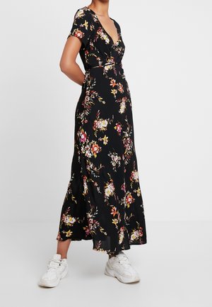 Kobieta w czarnej, kwiatowej, maxi sukience z krótkim rękawem, nosząca białe masywne sneakersy, stoi z rękami za plecami.
