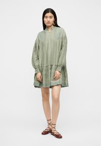 Γυναίκα που στέκεται φορώντας ένα χαλαρό, ανοιχτοπράσινο, μακρυμάνικο φόρεμα με δαντελένιες λεπτομέρειες και κολάρο με κουμπιά, συνδυασμένο με καφέ δερμάτινα παπούτσια με κορδόνια.