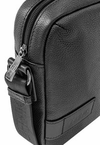 Czarna skórzana torba o teksturowanej powierzchni, zamek błyskawiczny, srebrne akcesoria oraz pleciony pasek z naszywką logo z przodu.