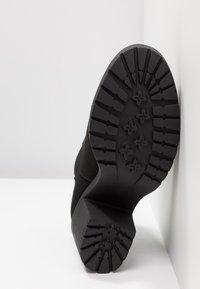 Semelle d'une botte noire avec des crampons profonds et un talon épais reposant contre un coin de mur blanc et de sol.