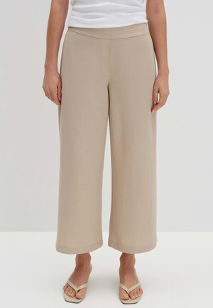 Kvinde iført beige vidde kortbukser og matchende beige sandaler, stående mod en ensfarvet lys baggrund.