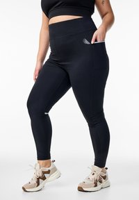 Czarne legginsy wysoki stan z bocznymi kieszeniami, o gładkiej fakturze i przeszywanych szwach. W połączeniu z beżowymi i brązowymi butami sportowymi.