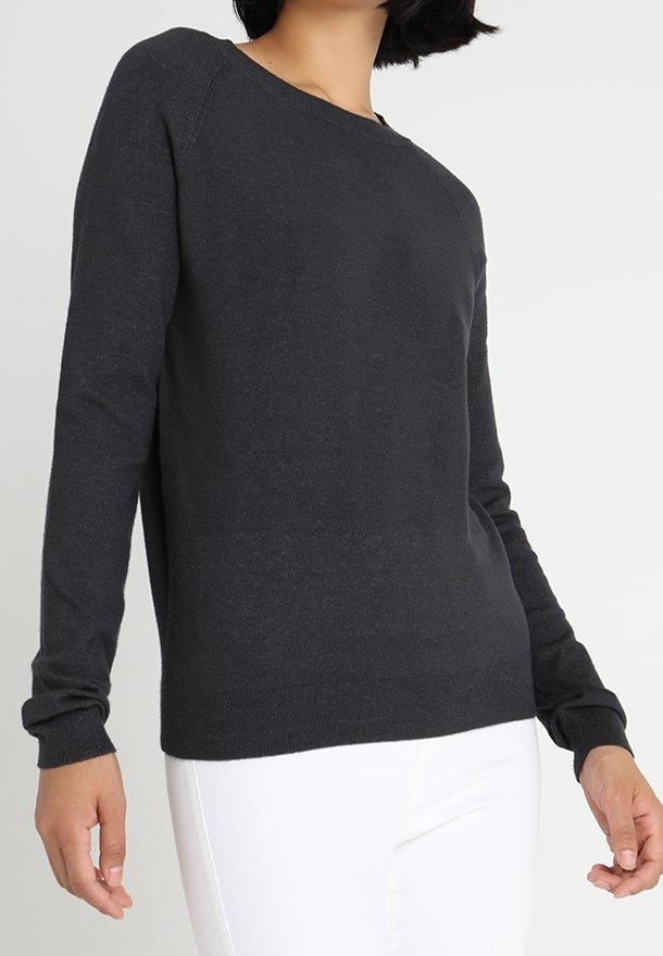 Femme portant un pull à manches longues gris foncé uni associé à un pantalon blanc, debout devant un fond clair uni.