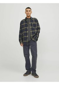 Camisa de franela a cuadros en azul marino y caqui, con bolsillo frontal, combinada con una camiseta gris y pantalones oscuros. Zapatillas negras con textura completan el atuendo.