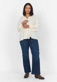 Blusa di un leggero avorio con fronte plissettato, maniche lunghe a palloncino e chiusura con bottoni, abbinata a jeans svasati blu scuro e stivaletti marroni.