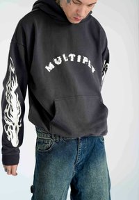 Sort hættetrøje med teksten "MULTIPLY" over brystet, flammer på ærmerne, parret med falmede blå denimjeans. Blød tekstur, afslappet design.