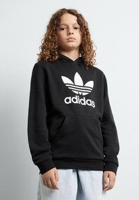 Fekete kapucnis pulóver nagy, fehér Adidas trefoil logóval. Elől zseb található, és húzózsinórral rendelkezik a kapucni. Simább anyagból készült, kényelmes fazonban.