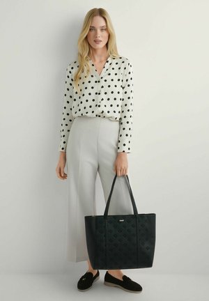 Kobieta o długich blond włosach nosi białą bluzkę w czarne kropki, jasnoszare spodnie, czarne mokasyny i trzyma dużą czarną torbę typu tote.