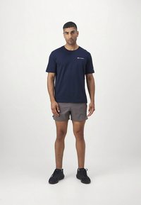 Tumši zils īsroku t-krekls ar baltu logotipu kreisajā krūtīs, kombinēts ar pelēkiem šortiem un melnām sporta kurpēm. Vienkāršs, ikdienišķs dizains.