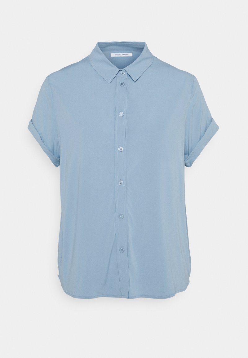 Chemise bleu clair à manches courtes avec boutons, manches retroussées et col pointu, présentée sur un fond blanc uni.