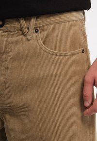 Brune kordfløjspants med lodret ribbet tekstur, der har en frontlomme og sorte metalaccenter ved taljen og lommen.