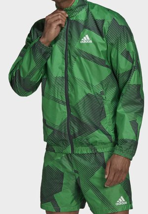 Man adjusting zipper on green Adidas jacket with black geometric patterns, matching shorts, both featuring white Adidas logos.