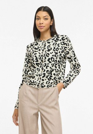 Femme portant un cardigan boutonné à imprimé léopard noir et blanc et un pantalon beige, debout avec une main dans la poche.