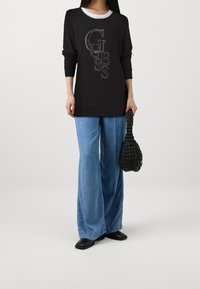 Pull noir oversize avec un logo "GUESS" métallique texturé. Associé à un jean large bleu clair et un sac à main tressé noir.