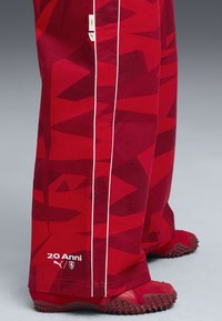 Rode broek met een geometrisch patroon, voorzien van witte zijstrepen. Accenten zijn het "20 Anni" logo en tekst nabij de enkel. Structuurstof.