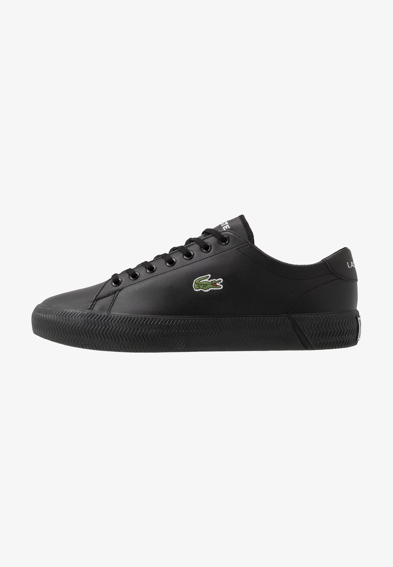 Baskets basses noires avec lacets noirs, semelle texturée et petit logo crocodile vert sur le côté.