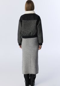 Vrouw met de rug naar de kijker, gekleed in een zwart-grijs herringbone jas met een witte kraag, een grijze gebreide rok en zwarte kniehoge laarzen op een witte achtergrond.