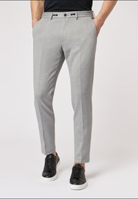 Graue maßgeschneiderte Hose mit glatter Textur, Slim-Fit und zwei Seitentaschen. Kombiniert mit schwarzen Sneakers mit weißen Sohlen.