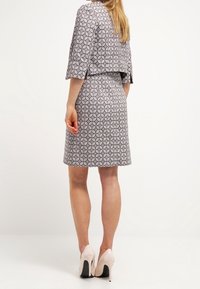 Kobieta stojąca tyłem, ubrana w sukienkę do kolan w wzory, z dopasowaną, krótką kurtką oraz jasnoróżowymi szpilkami.