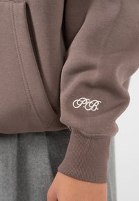 Manica di un sudario marrone con polsino a coste e iniziali ricamate in bianco "P.B.", mostrata sul braccio di una persona su uno sfondo di gonna grigia.