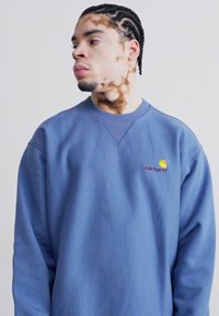 Blå sweatshirt med rund hals lavet af bomuld, præget af et lille Carhartt-logo i gul og mørk maroon på venstre bryst. Glat tekstur.