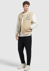 Jachetă varsity bej cu patch A, mâneci culoarea crem, nasturi negri și manșete cu dungi, asortată cu pantaloni negri și teniși albi.