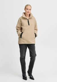 Beige fleece pullover med høj krave, halv lynlås, sorte detaljer og sidelommer, parret med mørkegrå bukser og hæleboots.