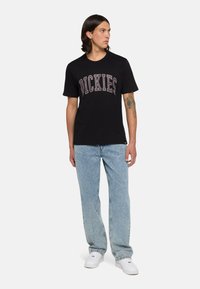 Camiseta de algodón negra con el texto destacado "DICKIES" en gris claro. Combinada con unos jeans de mezclilla azul claro y zapatillas blancas.