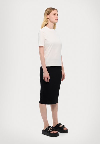 T-shirt blanc à manches courtes, jupe noire jusqu'aux genoux, portant des sandales à plateforme noires avec des boucles décoratives ; tissu en coton lisse, silhouette ajustée.