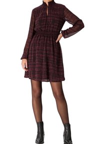 Ein rot-schwarz gemustertes Kleid mit hohem Kragen, langen Ärmeln, elastischem Taillenbund und lässigem Schnitt, kombiniert mit schwarzen Stiefeletten.