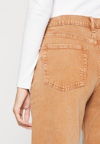 Jeans de tiro alto en color marrón mostrando el detalle del bolsillo trasero y las costuras, combinados con una blusa blanca de manga larga que resalta la suave textura de la tela.