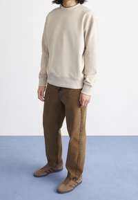 Beige sweatshirt med rund hals och ribbat avslut, parat med bruna jeansbyxor och bruna sneakers med mörka ränder.