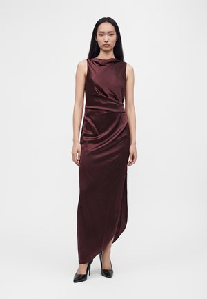 Femme aux longs cheveux noirs portant une robe en satin bordeaux foncé sans manches et des talons hauts noirs, debout devant un fond blanc.