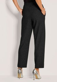 Vrouw in zwarte getextureerde wijde broek en zilveren hoge hakken staat met handen in haar zakken tegen een lichte muur.