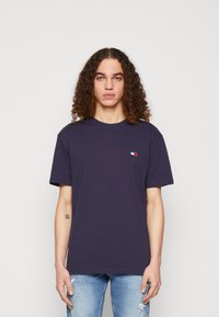 T-shirt in cotone navy con maniche corte, scollo a girocollo e una piccola toppa con logo in rosso, bianco e blu. Indossato con jeans chiari a effetto distressed.