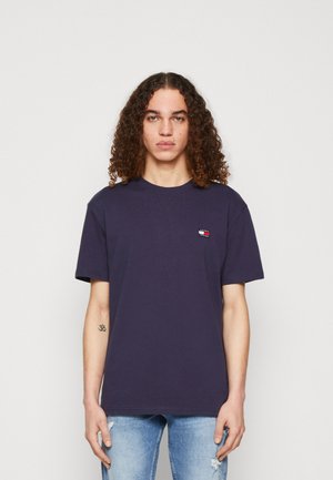 Camiseta de algodón azul marino con mangas cortas, cuello redondo y un pequeño parche de logo en rojo, blanco y azul. Llevada con jeans claros desgastados.