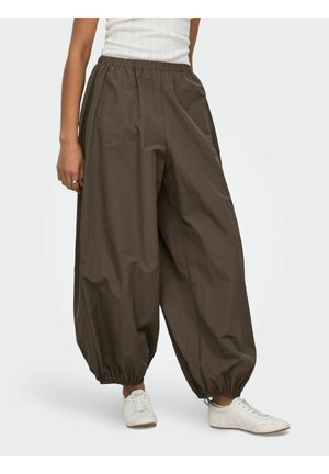 Personne portant un pantalon ample taille haute de couleur brun foncé avec des poignets élastiques, un haut blanc sans manches et des baskets blanches sur un fond clair.