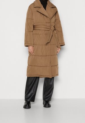 Personne portant un long manteau matelassé marron avec ceinture, un col roulé noir, un pantalon large noir et des chaussures épaisses noires, debout contre un mur blanc.