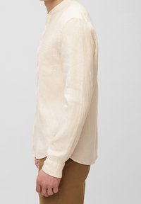 Uomo che indossa una camicia di lino beige a maniche lunghe con taschino e pantaloni marroni, in piedi contro uno sfondo semplice e chiaro.