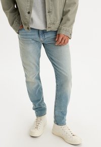 Unos vaqueros de mezclilla azul claro presentan un corte relajado con un efecto de desgaste, combinados con unas zapatillas blancas de tipo alto y una camiseta gris debajo de una chaqueta verde.