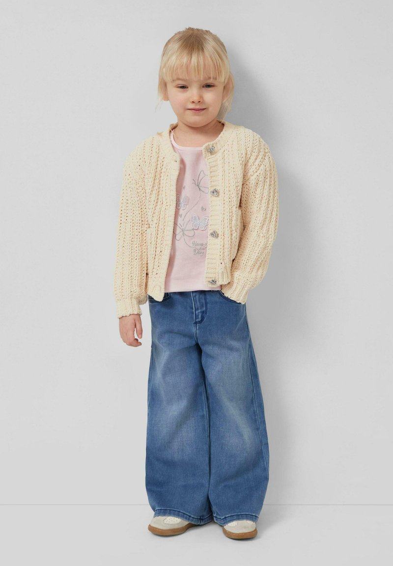 Mlada djevojka plave kose nosi kremastu pletenu vestu, ružičastu majicu s leptirima, široke traperice i bež tenisice, stojeći uz sivu zid.